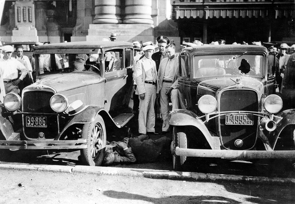 A crowd of onlookers gathers at the site of the Union Station Massacre of June 17, 1933. One of the fallen officers can be seen on the ground between the two automobiles.